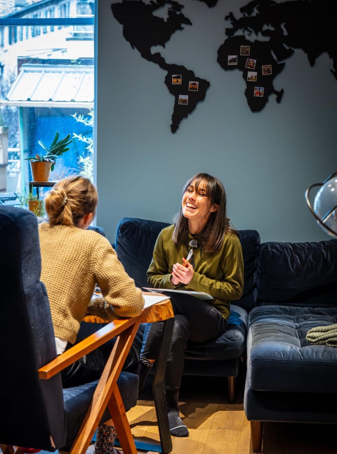 Nina Barrera en pleine interview, assise face à son invitée dans un salon lumineux, carte du monde au mur