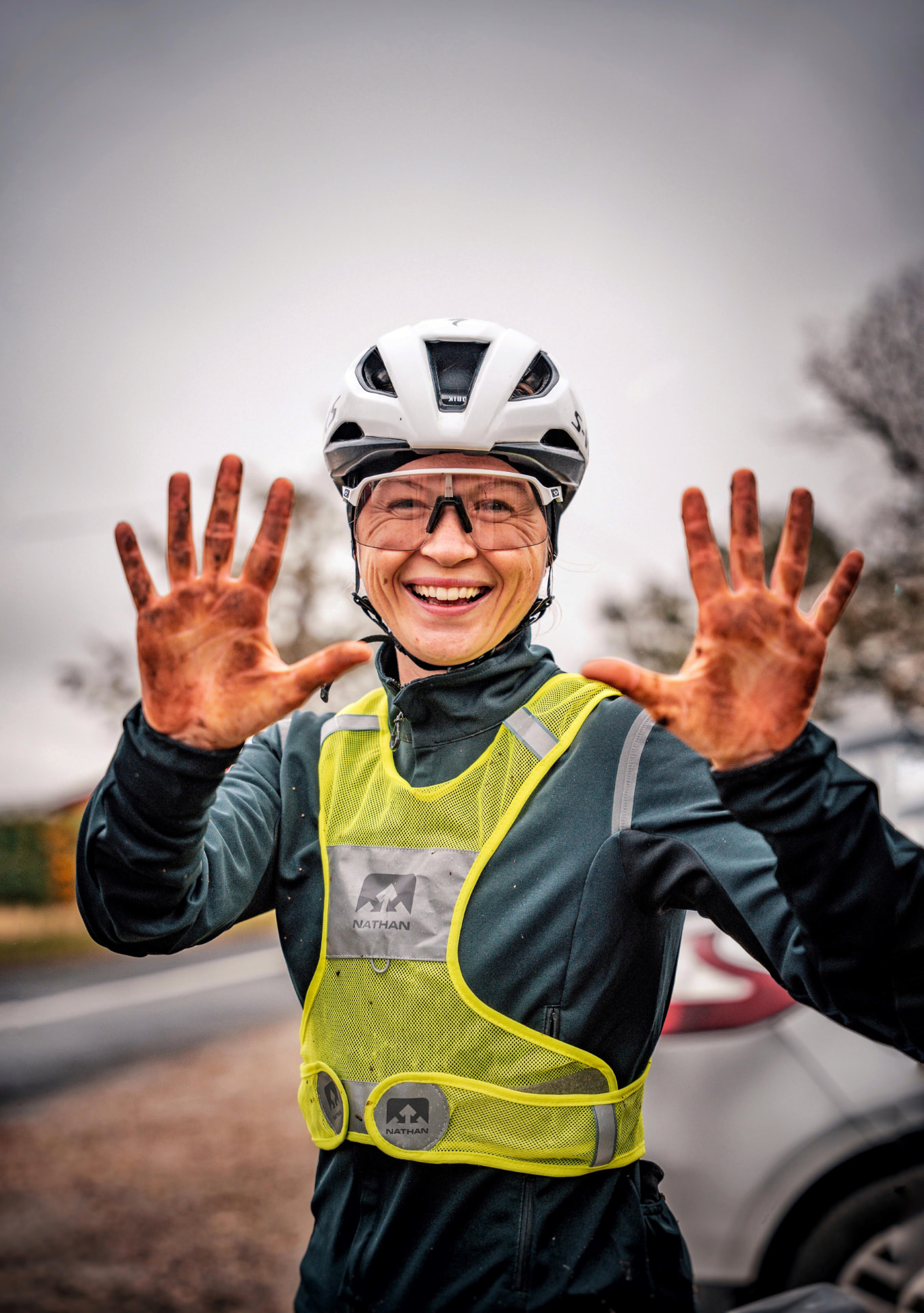 Cycliste souriante en gilet fluo montrant ses mains couvertes de boue après une sortie